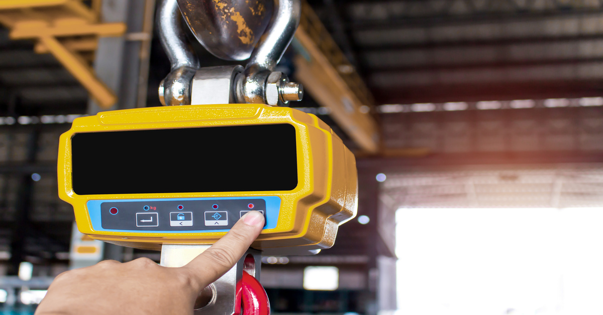A finger pressing a button on a yellow overhead crane scale inside a warehouse. The scale's digital display is blank.