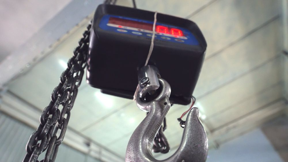 A crane scale with a digital display and large metal hook hanging from the ceiling inside an industrial environment.