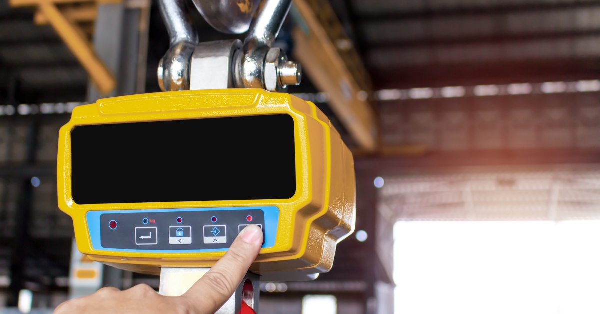 A hand pushes a button on a yellow digital crane scale hanging from a metal hook inside an industrial environment.