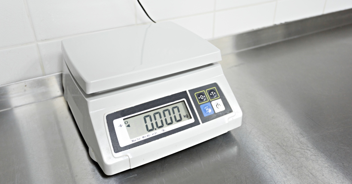 White scale with a digital display showing 0 with four buttons placed over a stainless steel table in front of a white wall.