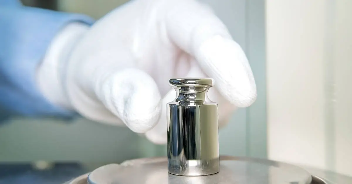 Close-up of a hand wearing a white glove placing a small metal weight on top of the flat metal surface of a scale.