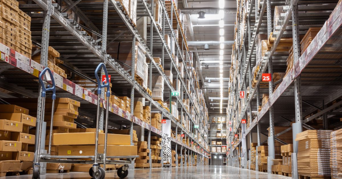 A well-lit warehouse aisle with tall shelves of cardboard boxes on either side and a flatbed cart in the center.