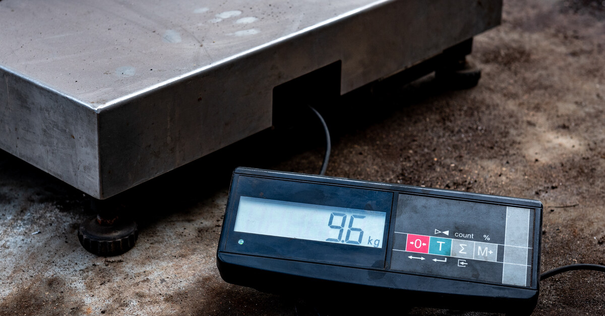 A silver platform scale rests on a concrete floor and weighs a pile of wires. An attached screen displays 9.6kg.