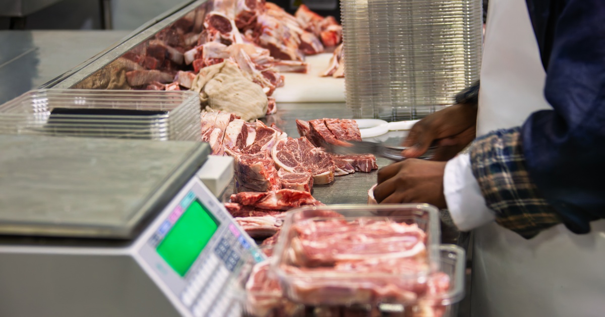 A worker in a white apron handling meat. He's at a table holding a digital scale, beef steaks, and plastic containers.