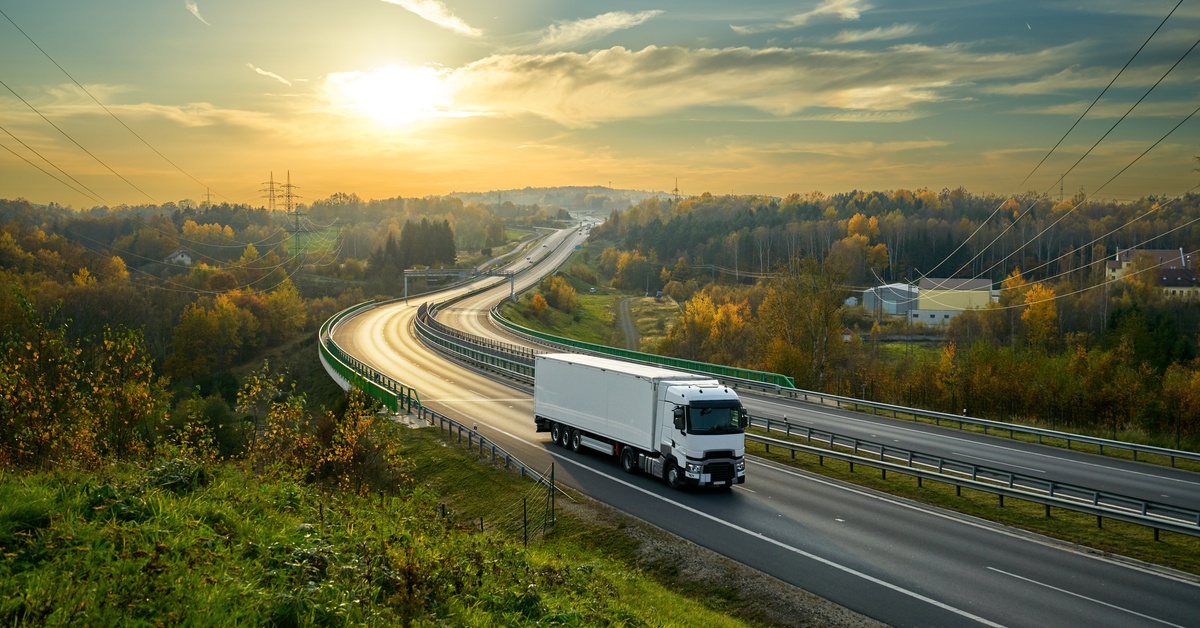 A white truck is driving on a highway, surrounded by forestry terrain and buildings on the right side in the distance.