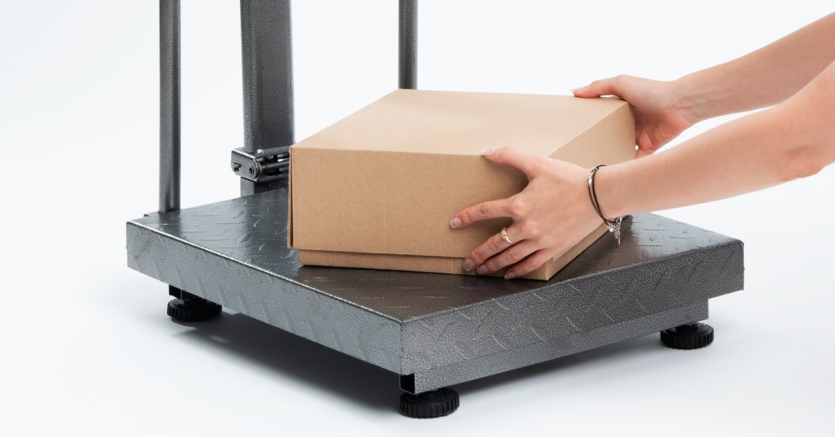 A pair of hands places a brown package on a weight scale. The scale has a screen with a white, yellow, and red keypad.