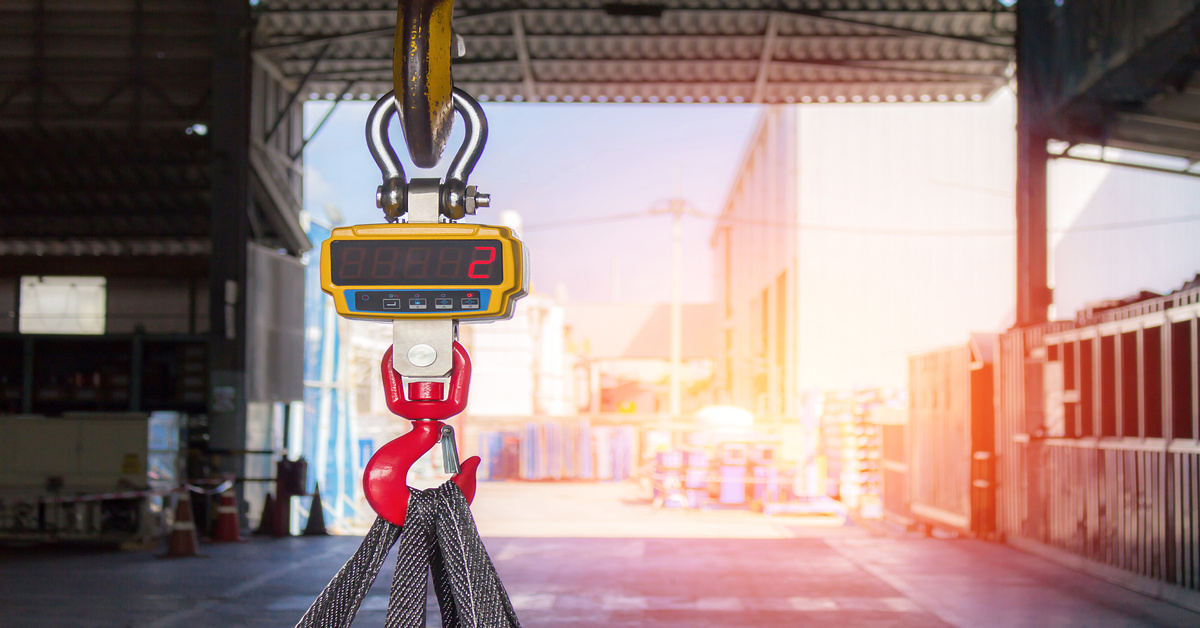 An industrial digital crane scale measures materials in an outdoor warehouse, with the meter displaying a reading of two.