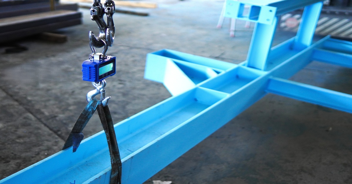 A digital crane scale with straps lifting a heavy blue steel beam, ensuring safe handling in an industrial setting.