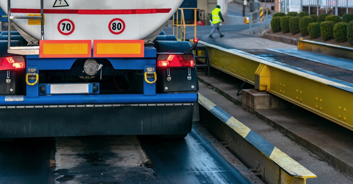 A close-up of a tank truck on a scale and the driver in the background walking taking the papers to the weighing control.