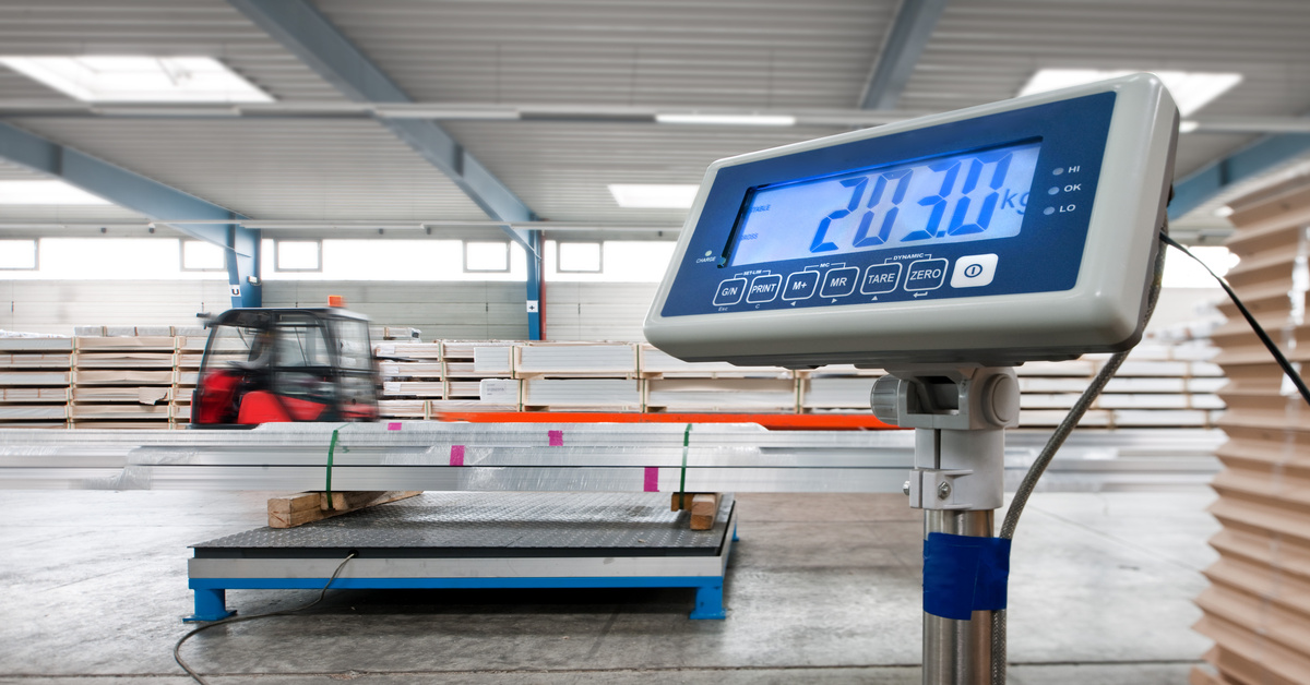 A close-up of the reader of an industrial floor scale with steel beams on it in a warehouse with a truck in the background.