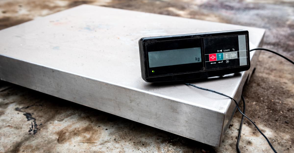 A silver commercial platform scale with a black digital reader attached. Both devices sit on a stone surface.