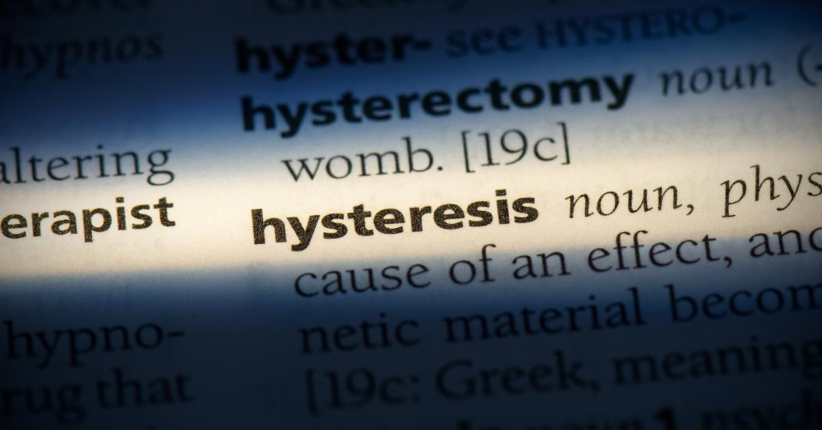 A close-up of a dictionary page highlighting "hysteresis" with warm light, the surrounding entries obscured in a dark blue shadow.