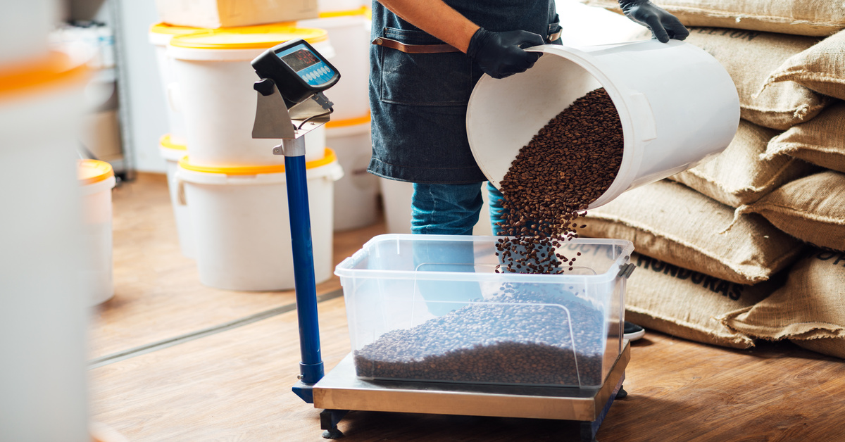 Someone pours a bucket of coffee beans into another container that is resting on a large weighing scale.