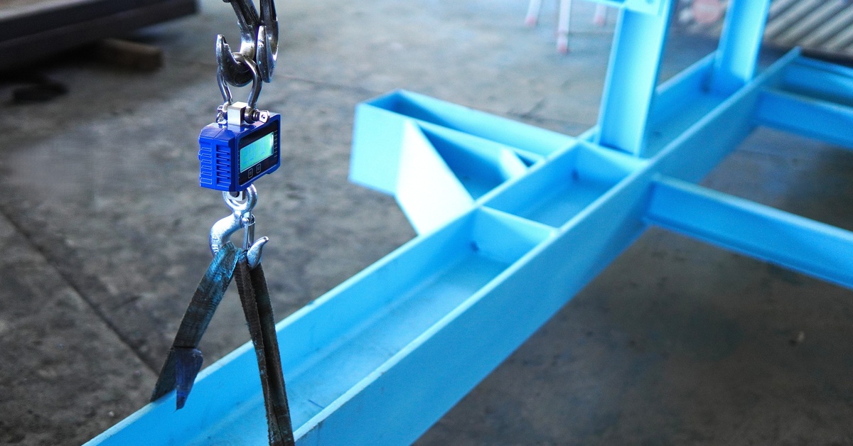 A blue crane scale attached to the long end of a blue-painted steel beam with a black strap and silver hook.