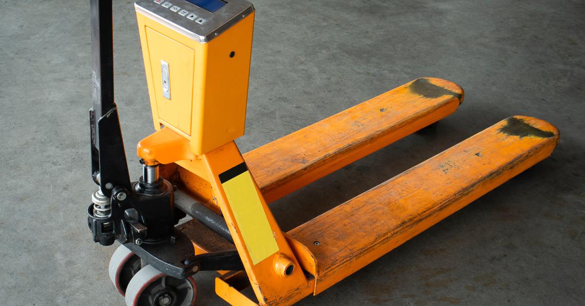 A close-up of an orange and black pallet jack with a built-in digital scale. The jack is parked on a concrete surface.