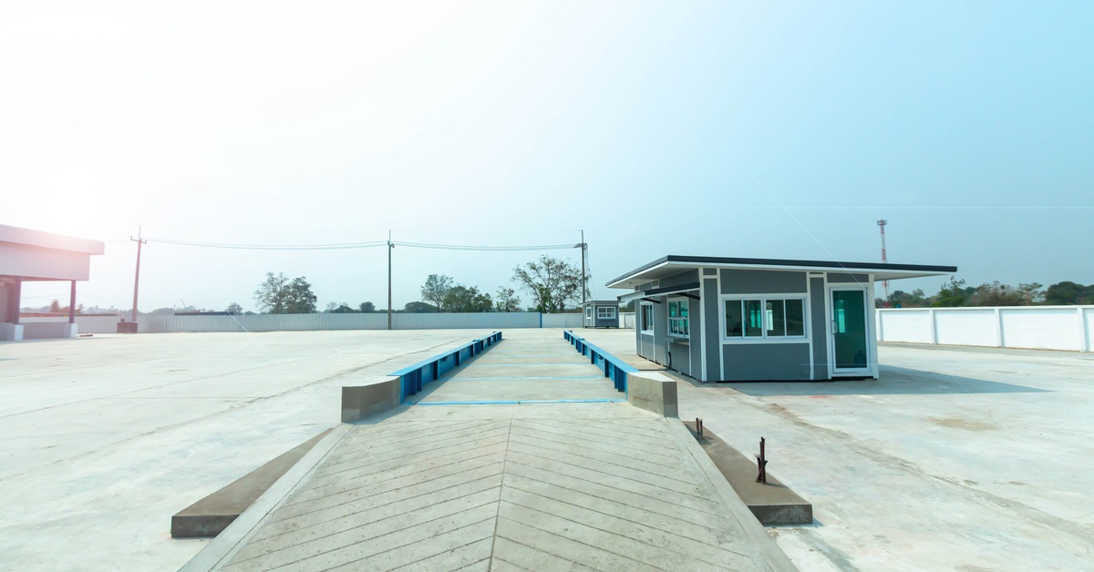 An elevated truck scale with blue side rails and concrete ramps set up next to a small building in an open, paved lot.
