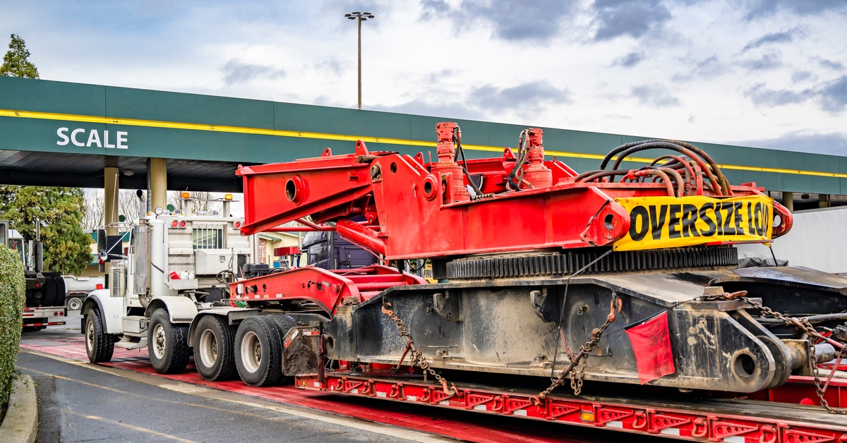 A white semi-truck hauling a trailer with an oversized load onto a set of red truck scales at a weighing station.