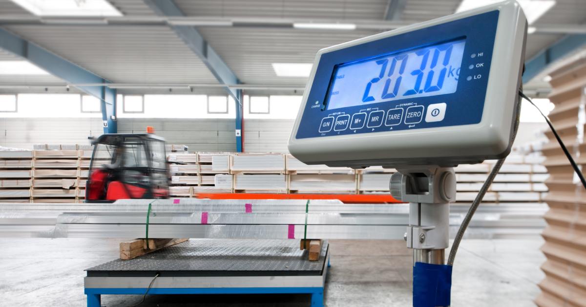 A close-up of the digital display for an industrial floor scale. The floor scale behind the display is holding metal beams.